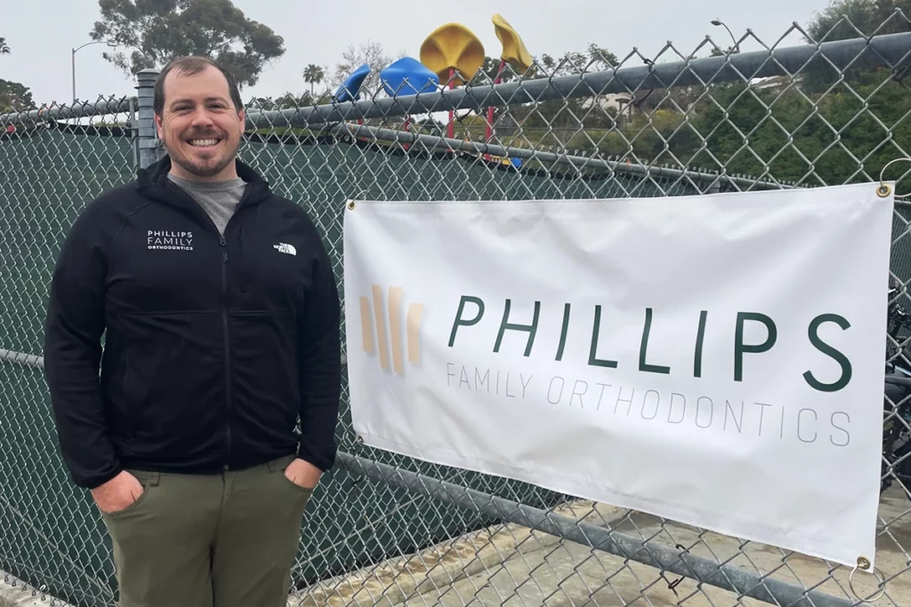 Dr. Joseph Phillips, orthodontist, is dedicated to community impact through partnerships and sponsorships, as seen next to the Phillips Family Orthodontics banner supporting local elementary schools in Mira Mesa and Encinitas, CA.